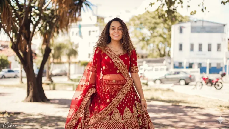 A young woman in a rich, royal red bridal lehenga. The blouse and the full, voluminous flared skirt feature intricate gold zari embroidery, kundan stone work, and detailed traditional motifs. A sheer red dupatta with heavy embroidered borders is draped over her left shoulder and head, with her right arm and hand holding the dupatta in a different pose than previous images, showcasing the fabric's heavy texture. She wears a small necklace. She stands in a sun-drenched park with palm trees, blurry buildings, and vehicles in the background. Her face, curly hair, and features are clearly visible and identical to the original subject. A young woman in a rich, royal red bridal lehenga. The blouse and the full, voluminous flared skirt feature intricate gold zari embroidery, kundan stone work, and detailed traditional motifs. A sheer red dupatta with heavy embroidered borders is draped over her left shoulder and head, with her right arm and hand holding the dupatta in a different pose than previous images, showcasing the fabric's heavy texture. She wears a small necklace. She stands in a sun-drenched park with palm trees, blurry buildings, and vehicles in the background. Her face, curly hair, and features are clearly visible and identical to the original subject.