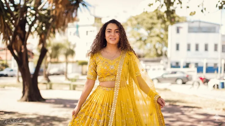 A young woman in a traditional bright yellow and gold lehenga. The blouse and long flared skirt are rich with gold thread embroidery, sequins, and gold embellishments. A sheer net dupatta with gold borders and motifs is draped over her left shoulder and head, held slightly forward. She stands in a sun-drenched park with palm trees, blurry buildings, and vehicles in the background. Her face, curly hair, and pose are clearly visible and identical to the original subject. A young woman in a traditional bright yellow and gold lehenga. The blouse and long flared skirt are rich with gold thread embroidery, sequins, and gold embellishments. A sheer net dupatta with gold borders and motifs is draped over her left shoulder and head, held slightly forward. She stands in a sun-drenched park with palm trees, blurry buildings, and vehicles in the background. Her face, curly hair, and pose are clearly visible and identical to the original subject.