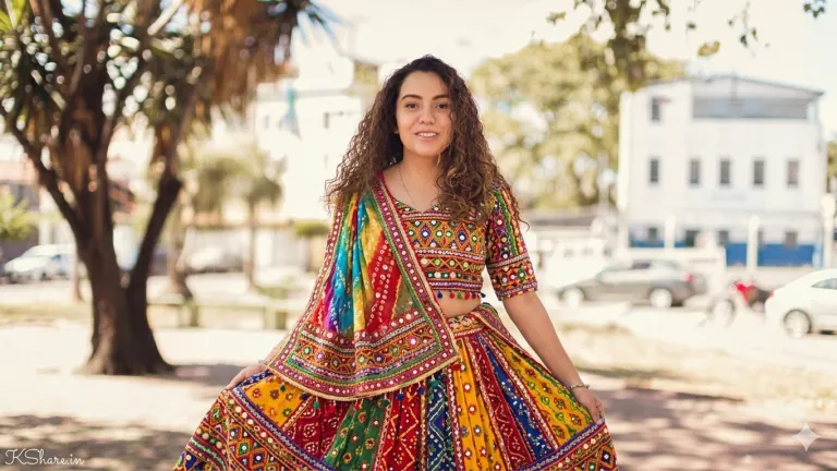 A young woman in a vibrant, multicolored Rajasthani lehenga. The full outfit, including the blouse, long flared skirt, and a draped dupatta, is densely covered in traditional embroidery and small, reflective mirror work in a 'kutch' style, creating a detailed patchwork of colors (red, yellow, green, blue). She stands in a sun-drenched park with palm trees, blurry buildings, and vehicles in the background. Her face, curly hair, and pose are clearly visible and identical to the original subject. A young woman in a vibrant, multicolored Rajasthani lehenga. The full outfit, including the blouse, long flared skirt, and a draped dupatta, is densely covered in traditional embroidery and small, reflective mirror work in a 'kutch' style, creating a detailed patchwork of colors (red, yellow, green, blue). She stands in a sun-drenched park with palm trees, blurry buildings, and vehicles in the background. Her face, curly hair, and pose are clearly visible and identical to the original subject.